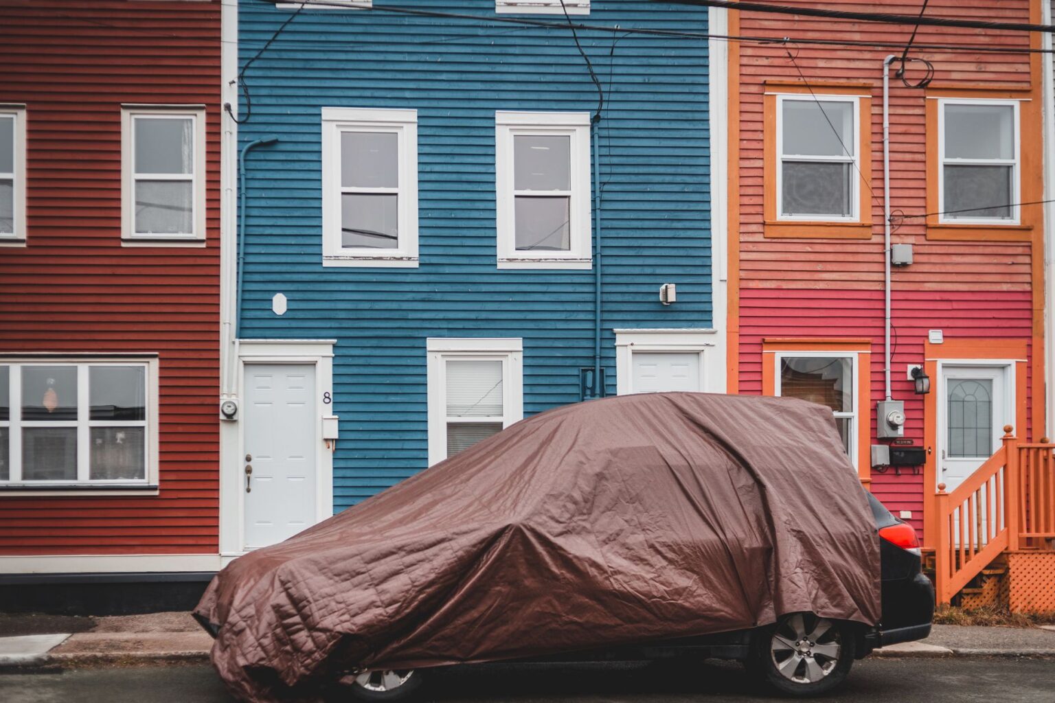 Automobile covered with brown fabric parked on road near colorful residential houses with windows and doors on street of town