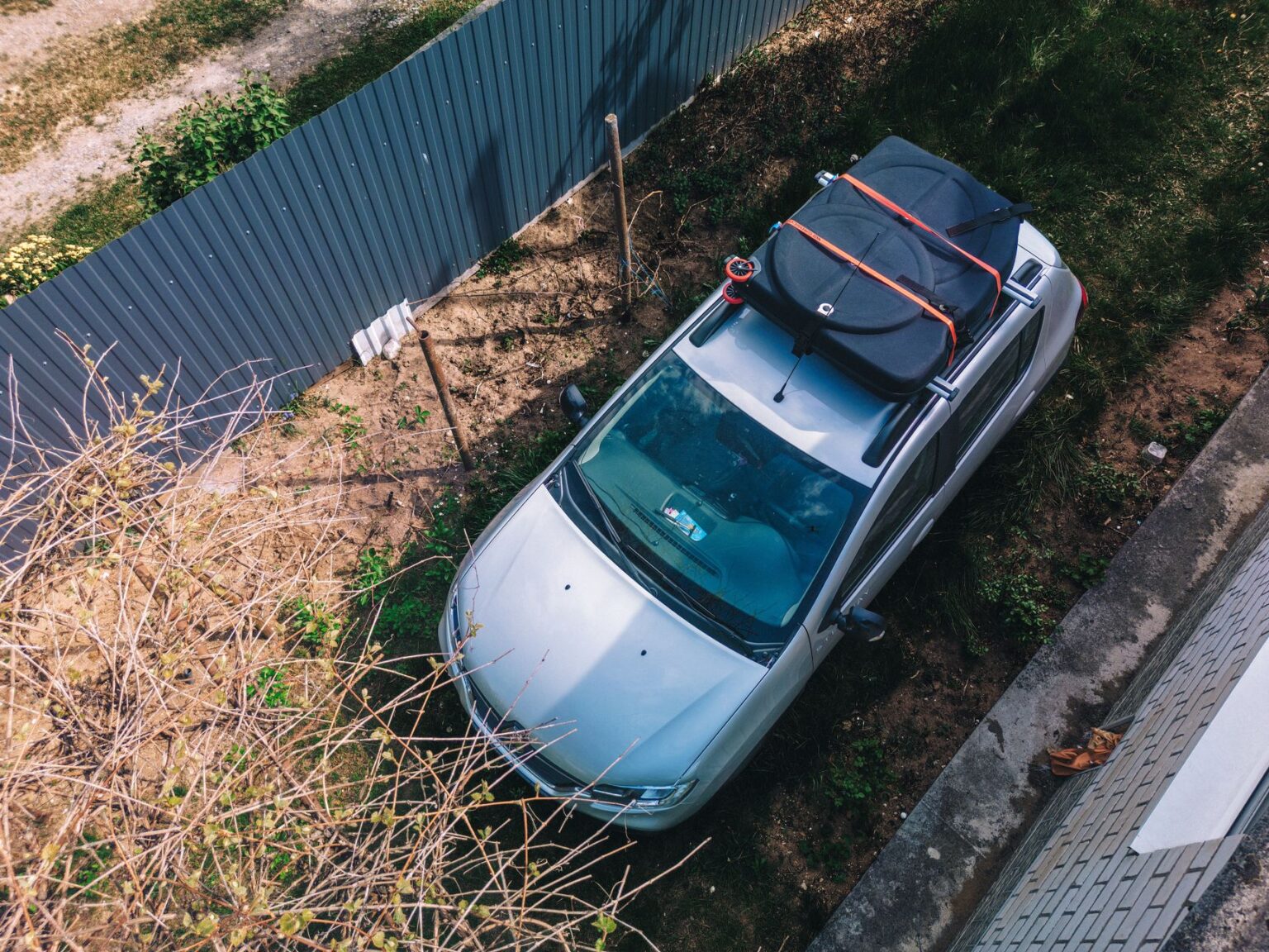 Aerial view of a silver car parked with a roof cargo carrier in a garden setting under clear daylight.