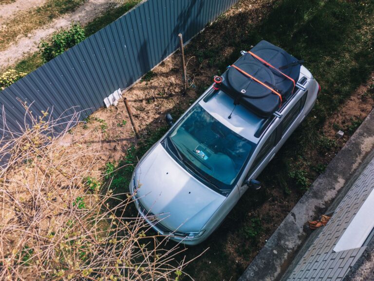 Aerial view of a silver car parked with a roof cargo carrier in a garden setting under clear daylight.