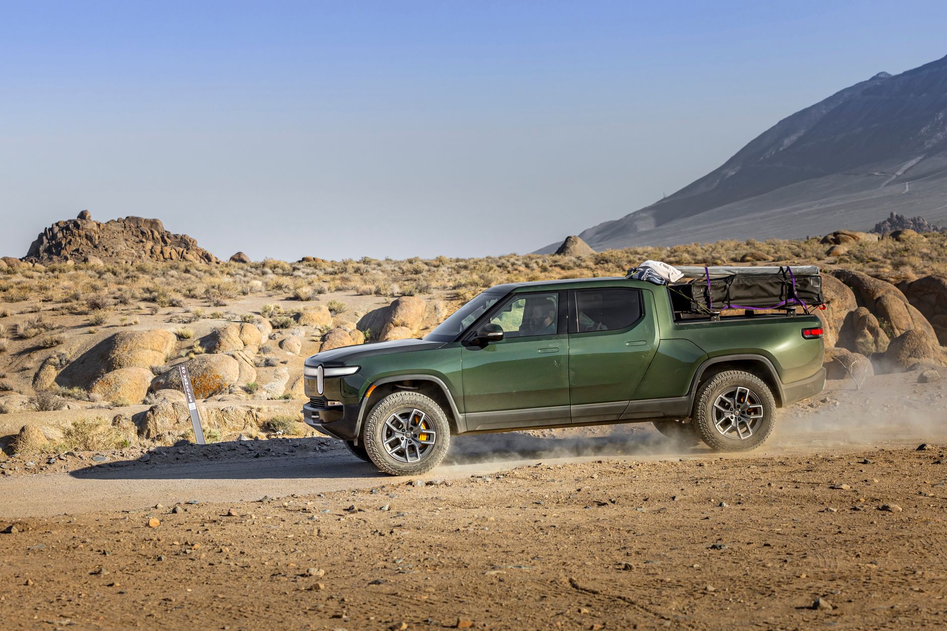 Green electric pickup truck on a dusty trail in a rocky desert landscape.