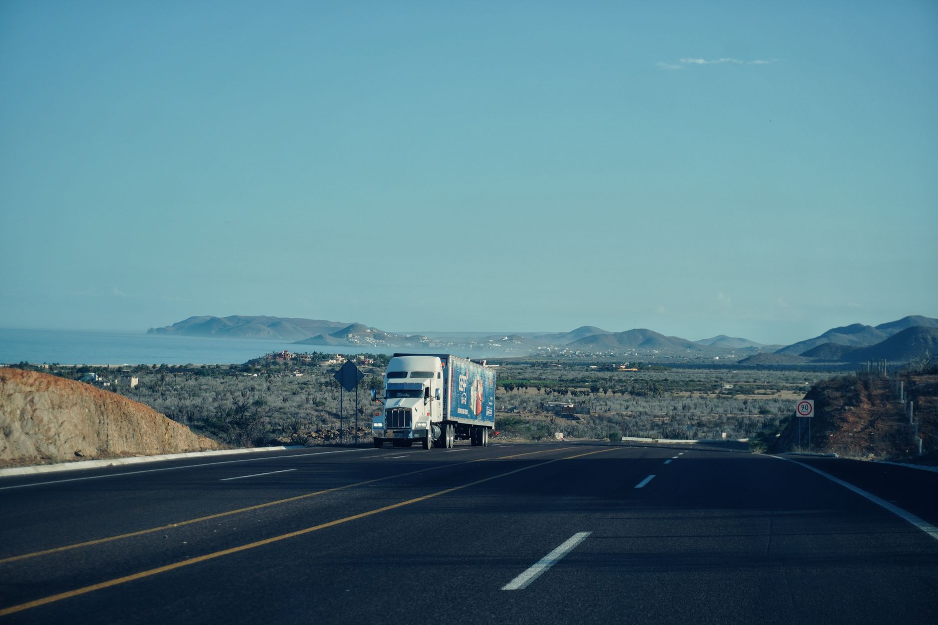 A semi-truck travels on a highway with mountains in the background.