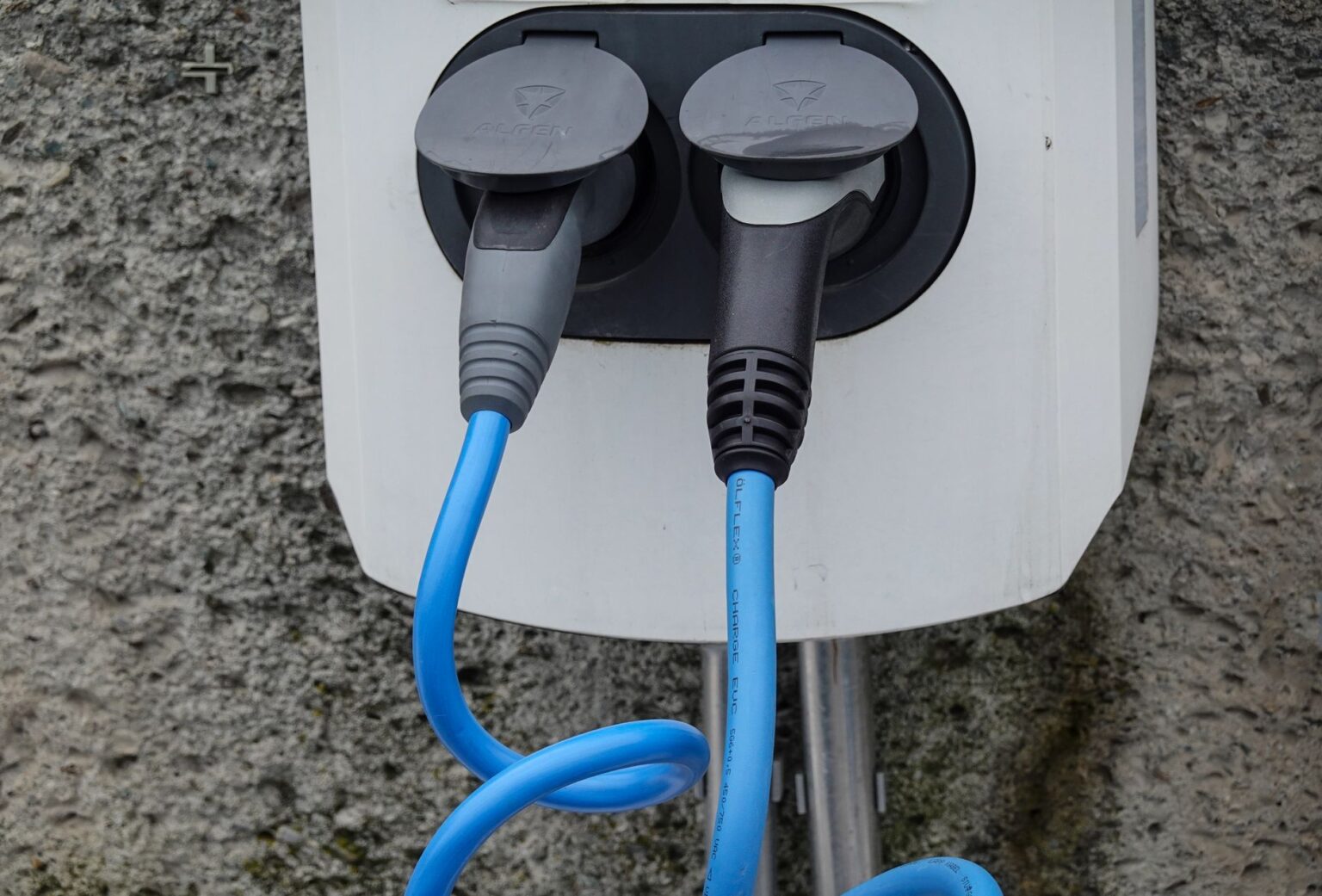 Close-up of an electric vehicle charging station with two cables in sockets against a textured wall.