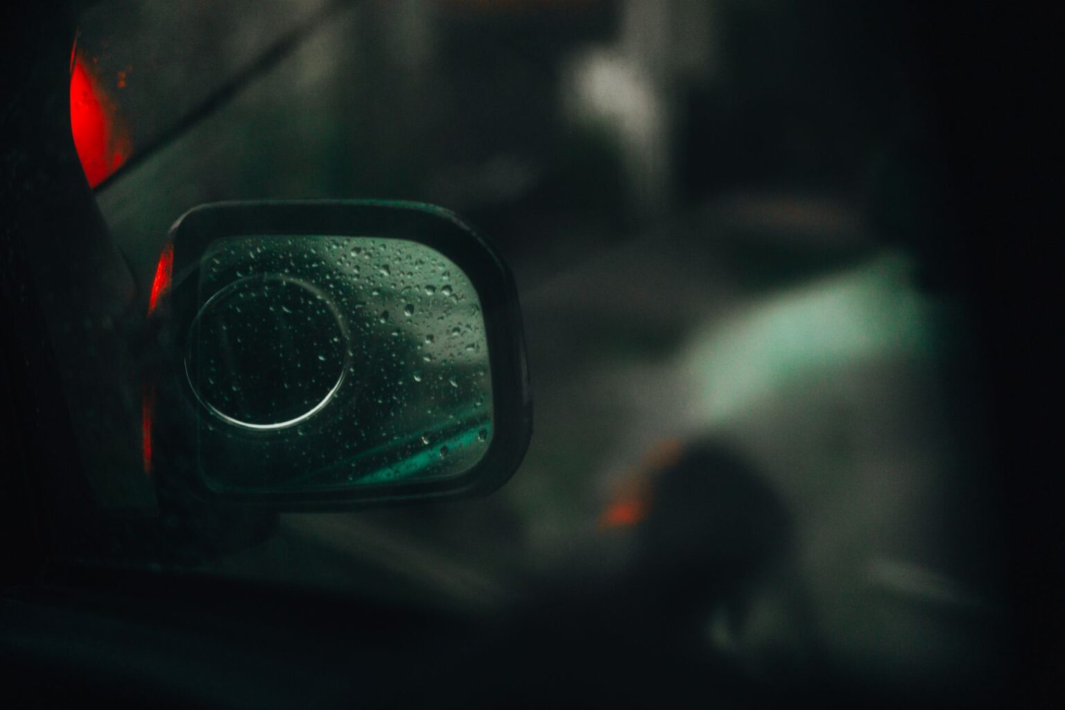 A close-up of a car's side mirror with raindrops at night, highlighting the reflective surface and red tail light.