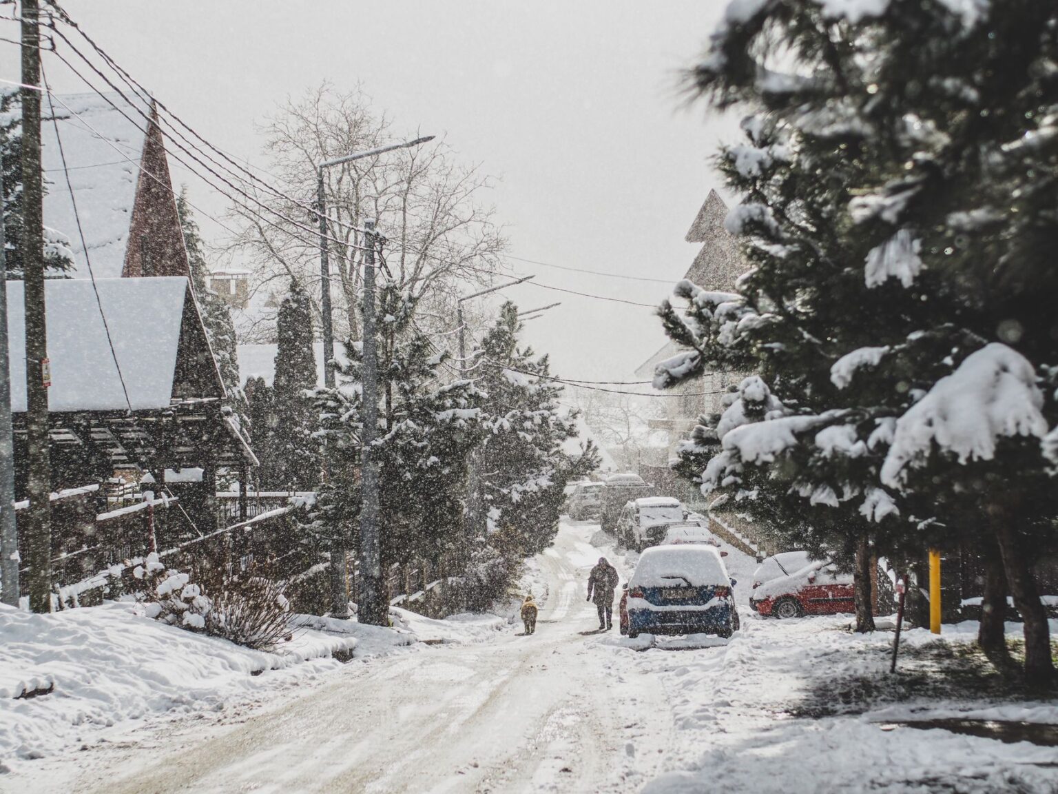 A winter scene of a snow-covered road with cars and people in a small town.